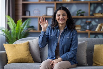 Portrait of a young smiling Indian woman in casual outfit sitting on the couch at home, waving and...