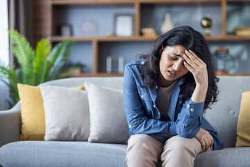 Tired young Indian woman in denim shirt sitting on sofa at home and holding her head, feeling...