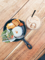 A delicious meal featuring rice, fried chicken, tempeh, fresh vegetables, and a refreshing iced coffee drink. The food is served in a skillet on a wooden tab