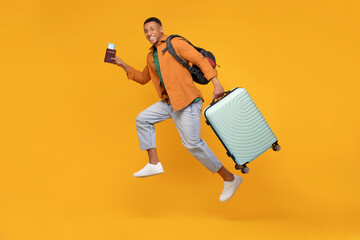 Happy African American man jumping with suitcase and passport, having fun over orange background,...