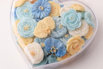 heart shaped container filled with colorful, intricately designed flower shaped soaps in shades of blue, yellow, and white, with gold accents, creating delicate and elegant appearance
