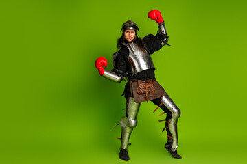Fototapeta premium Young man dressed as medieval knight with boxing gloves in a humorous representation of past and modern era, isolated on a green background