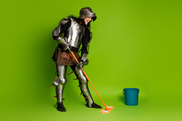 Young man dressed as a medieval knight cleaning the floor on a green background