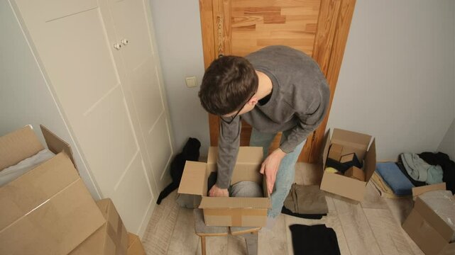 A man with black cat places folded clothes into an open cardboard box while preparing for a move. Surrounding him are multiple boxes and scattered clothes. He puts the cat in a box with his belongings