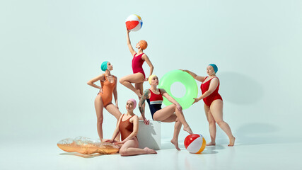 Five stylish women in vibrant swimsuits caps posing dynamically with float rings and beach balls against light blue background. Concept of summer, retro style, fashion, swimsuit, holiday, vacation