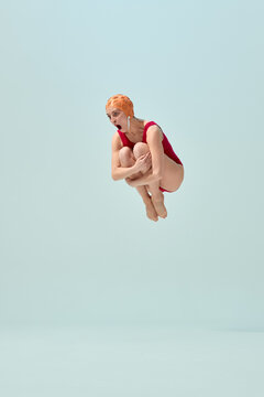 Diver in red vintage swimsuit and orange cap making cannonball pose in air with comic expression. Concept of summer fun, playful holiday, vintage swimsuit style, humor, retro sport.