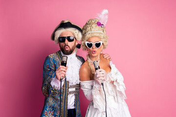 Elegant couple in vintage costumes singing karaoke under pink background at a theme party.