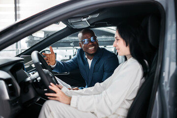 Fototapeta premium African-American dealer counseling a customer on buying a modern car at a car dealership. Concept of buying, leasing, test driving a new modern car. 