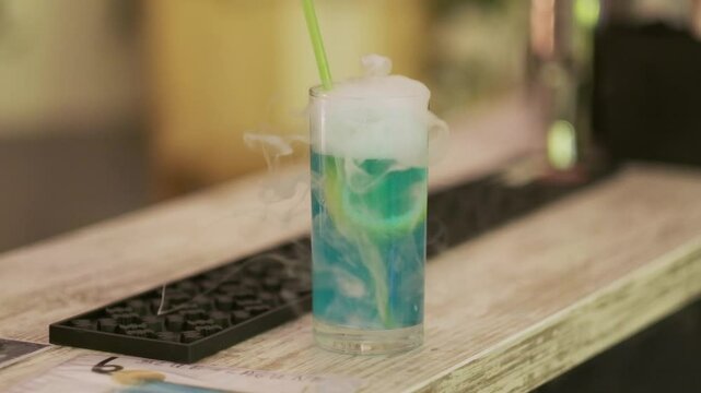 Blue cocktail with lemon slice served in a tall glass with smoke effect on a rustic bar counter


