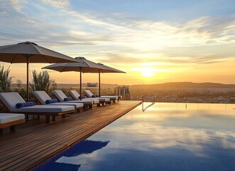 Photo of a modern swimming pool with a wooden deck and sun loungers under umbrellas at sunset in a hotel resort. The outdoor scenery features a city view in the background. 