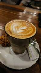 Portrait of love-shaped cappuccino coffee in a glass cup with white saucer and biscuits on a wooden table background.