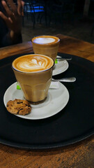 Portrait of love-shaped cappuccino coffee in a glass cup with white saucer and biscuits on a wooden table background.