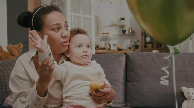Cheerful mother pushing colourful balloons with hand of baby daughter who eating apple on couch in living room