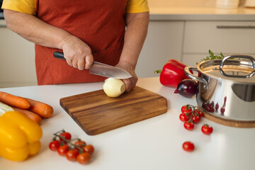 A woman in a yellow shirt and apron is peeling an onion in the kitchen. She is preparing ingredients for a meal while surrounded by fresh vegetables like carrots, tomatoes, and bell peppers