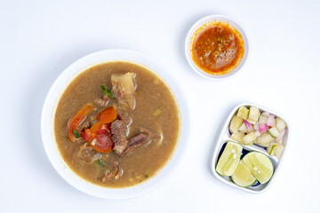 Overhead view of Soto daging sapi, a hearty Indonesian beef soup, in a white bowl. Accompanied by pickles, lime, sambal, and spoons on a clean, white surface.