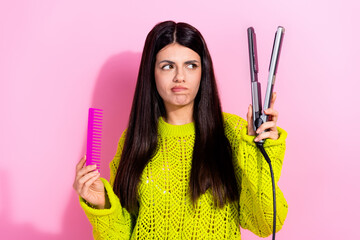 Smiling young woman debating hair styling options while holding a comb and flat iron against a...