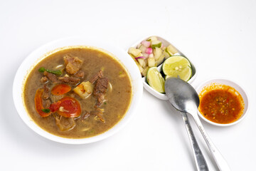 Overhead view of Soto daging sapi, a hearty Indonesian beef soup, in a white bowl. Accompanied by pickles, lime, sambal, and spoons on a clean, white surface.