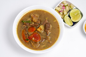 Overhead view of Soto daging sapi, a hearty Indonesian beef soup, in a white bowl. Accompanied by pickles, lime, sambal, and spoons on a clean, white surface.