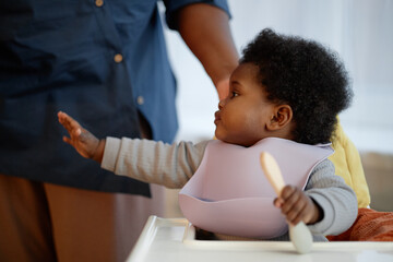 Side view of cute Black baby girl wearing silicone bib reaching out to take tasty dish during dinner time, while mother feeding little kid in kitchen