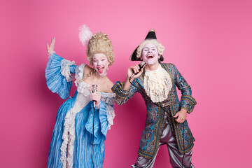 Fototapeta premium Young couple in extravagant baroque costumes joyfully posing against a vibrant pink background, embodying the elegance and charm