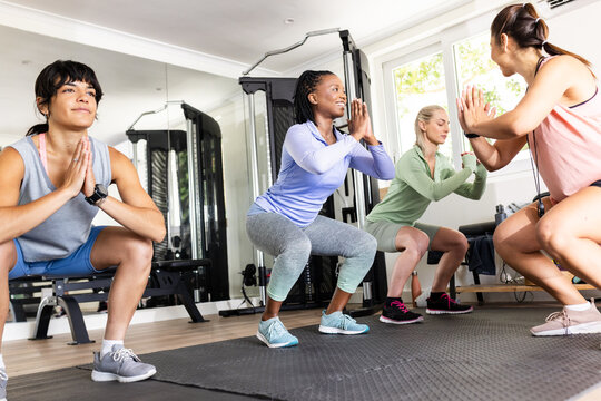 Diverse female friends exercising in gym, doing squats and smiling, promoting fitness, at home