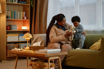 Affectionate Black mother playing with cute little baby showing lovely teddy bear to girl while...