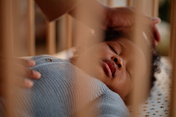 Shot through wooden crib slats of Black baby girl peacefully sleeping in bed while caring mother...