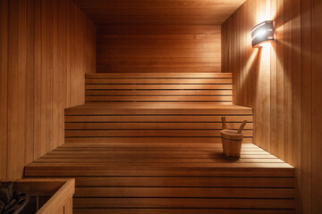 A designed wooden sauna with warm ambient lighting, featuring clean lines and a minimalist aesthetic. The space is furnished with tiered wooden benches and a traditional sauna bucket.