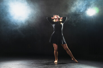 Contemporary woman dancer posing on black background with smoke. Adult female artist performer dancing with expression. Modern ballerina dancing in darkness. Passion dance in dark. Flexibility balance