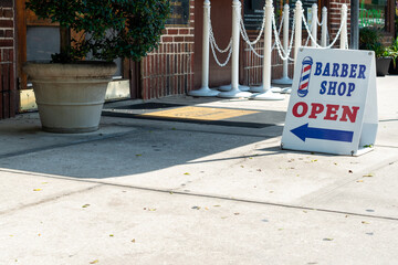 A white wooden sign on a sidewalk with a barber shop open and a blue arrow pointing to the entrance...