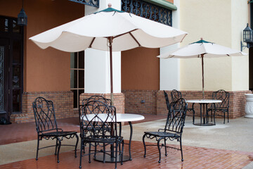 Multiple round white tables with large cloth sunshades and black metal bistro chairs surround the dining area, which is surrounded by large brick buildings, a concrete floor, and a stucco building.