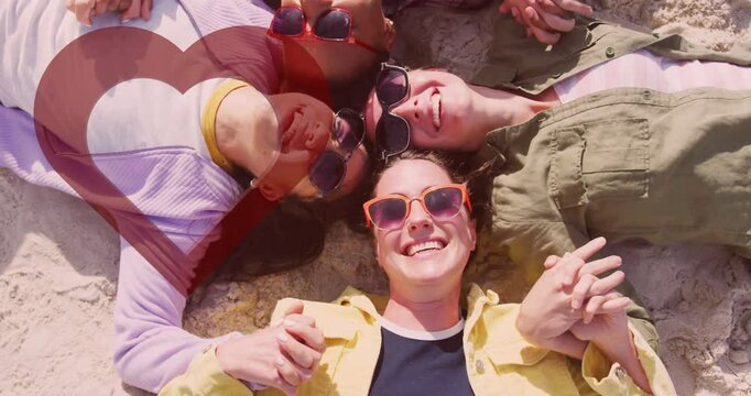 Wearing sunglasses, diverse friends lying on sand holding hands, smiling together