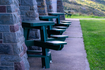 A row of green wooden pub-size outdoor tables and seats. The seating is against an exterior rock wall. There's green grass lawn and a view of hills and ocean in the distance. The wall is green and red