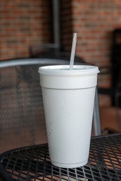 A large or tall white non-recyclable to-go drink cup with a plastic lid and straw on a black metal patio table. The polystyrene or Styrofoam insulated drinking cup is filled with a cold soda pop drink