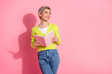 Young cheerful woman holding a book, wearing casual jeans and a yellow cardigan, posing against a vibrant pink background