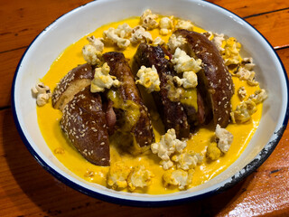 Unusual burger with pretzel bun, cheese sauce, bacon, and popcorn topping, served in a bowl. Creative street food presentation on rustic wooden table.