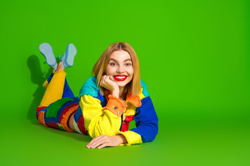 Colorful young woman smiling happily while lying on the floor against a bright green background in vibrant clothing fashion