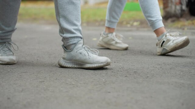 Lower angle view of two individuals walking side by side in grey joggers and sneakers, captured mid-step on paved road with greenery in blurred background