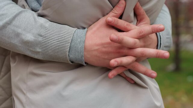 Close up of lovers embracing tightly around waist, hand resting gently on jacket, showing affection and intimacy, with soft background featuring blurred figure passing by on scooter