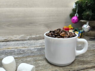 white cup filled with coffee beans next to pieces of marshmallows. There is a Christmas tree in the background