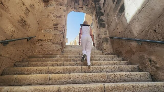 Woman wearing a hat and white outfit walks up stone stairs towards an archway in an ancient amphitheater in El Jem, Tunisia