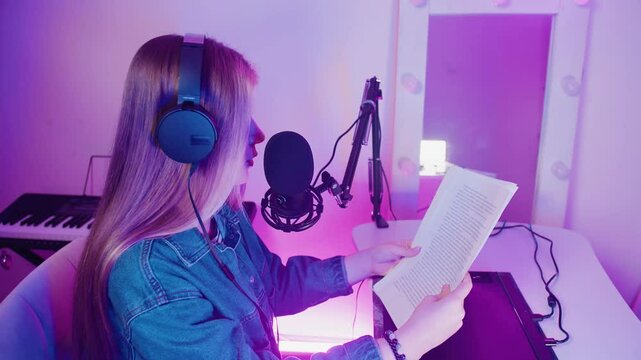 Side view of a woman in a denim shirt, with long hair and black headphones, sitting at a table and reading text from a piece of paper. Voice recording on a microphone in a purple professional studio