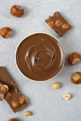 Chocolate spread in bowl, chocolate and hazelnuts on grey background, top view.