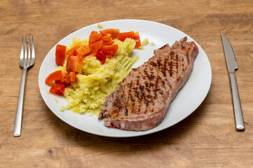 a steak, a beef entrecote on a white plate with mashed potatoes and chopped tomatoes on a wood table with a fork and a meat kife