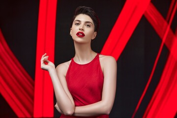 Stylish young woman with short hair and red lipstick poses in front of a striking red and black artistic background. Fashion, cosmetics, beauty editorial, or luxury branding concept.