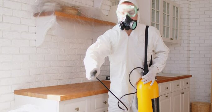 Worker in white protective suit, mask and gas respirator spraying insecticide in kitchen. Pest control specialist using professional equipment to eliminate bugs, cockroaches, insects or harmful pests.