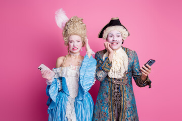 Couple dressed in baroque costumes with mobile phones against a pink background, showcasing a mix of historic elegance