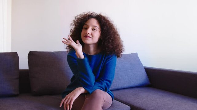 A young Hispanic woman with curly brown hair and a blue sweater smiles mischievously, then waves and teases playfully, adding a touch of flirtation to her expression