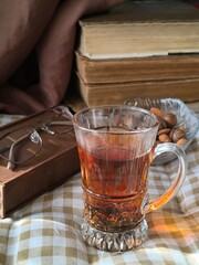 glass transparent cup of black tea and transparent plate with nuts, glasses in brown frame on wooden box against background of stack of books. brown background