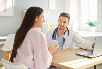 Obraz premium Smiling male doctor in white medical uniform meets to show checkup results on laptop, consult female patient, hand shaking, male GP happy to talk with woman client at meeting in modern hospital